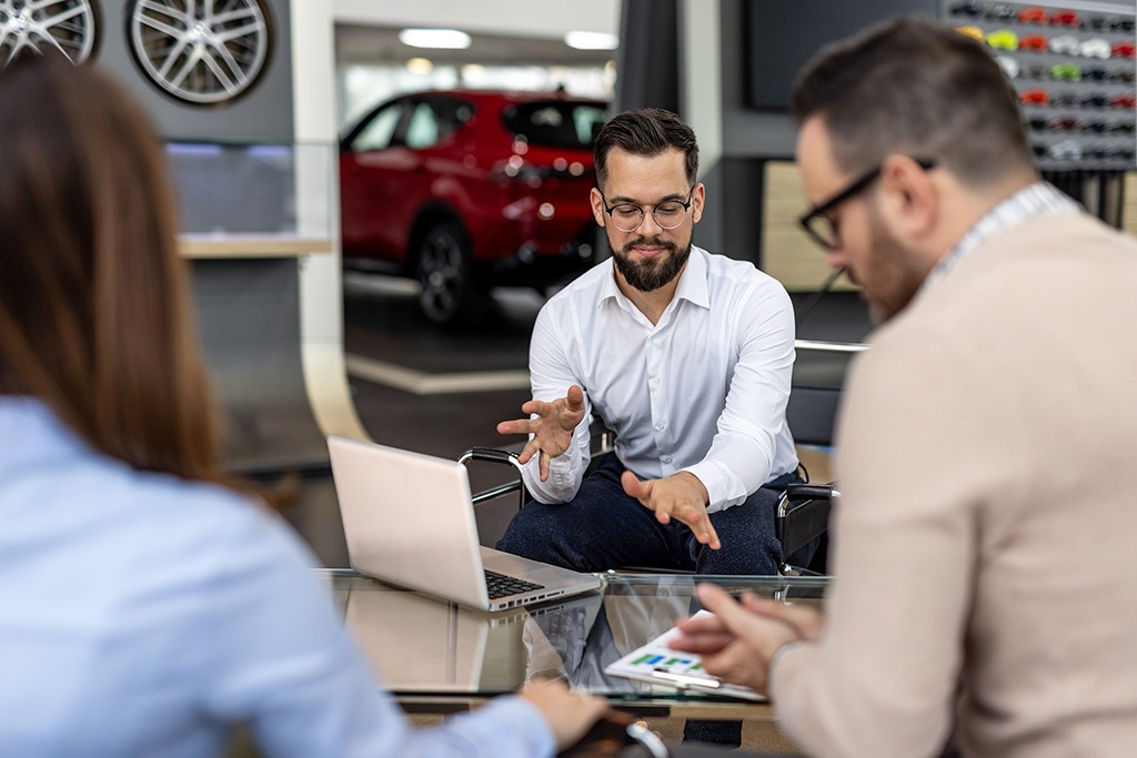A man in a white shirt and glasses gestures while explaining something to two colleagues seated around a glass table with a laptop and tablet. They appear to be in a modern car showroom, with a red vehicle and display of wheel rims visible in the background.