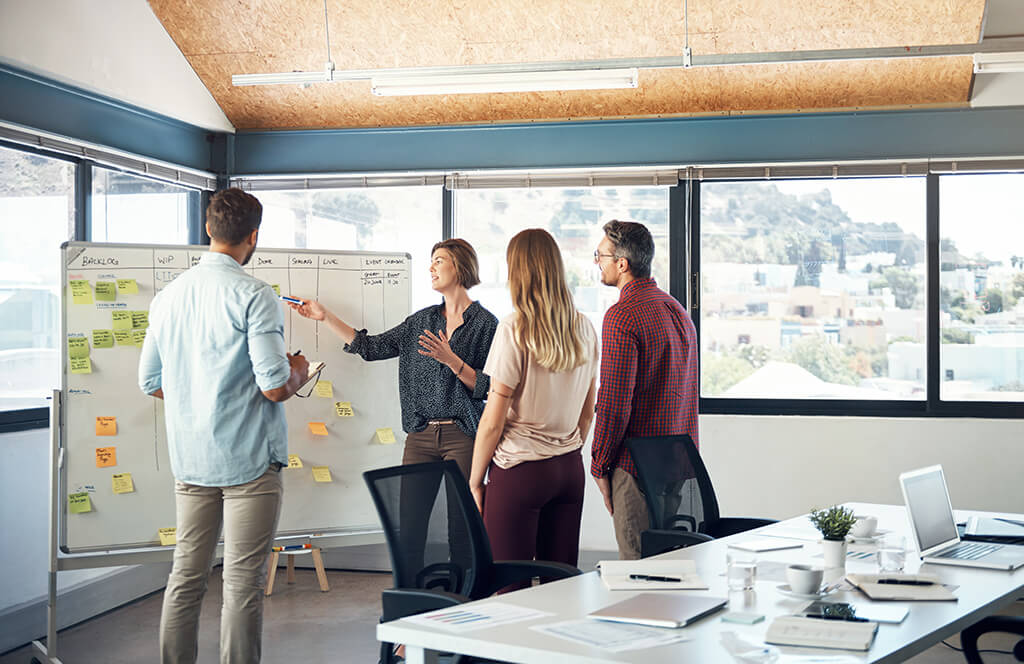 A woman leads a team meeting in a bright office, pointing to a whiteboard covered with sticky notes and project stages while three colleagues listen and discuss the plan.