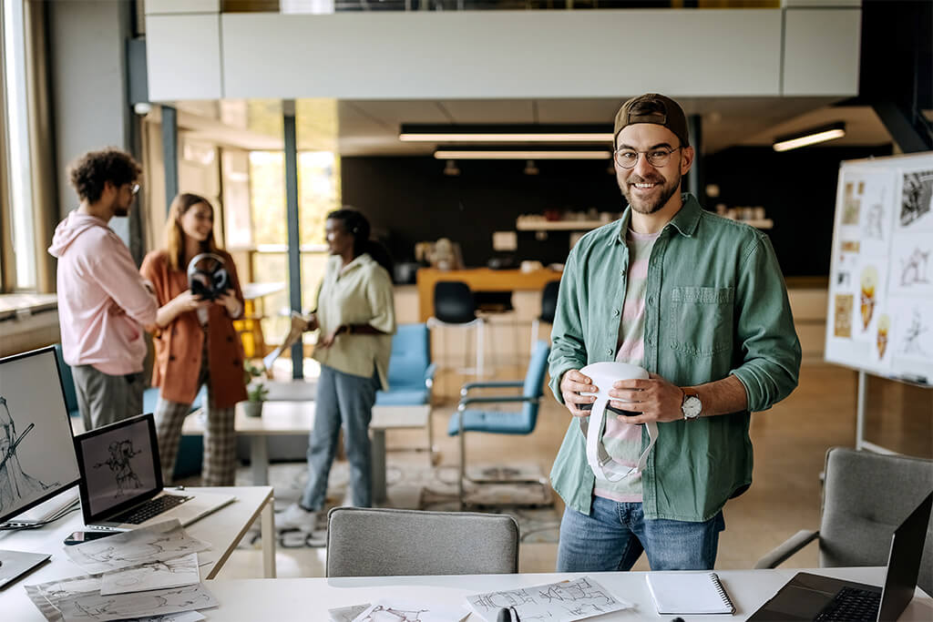 A smiling man wearing glasses and a green shirt holds a VR headset in a modern creative studio, with sketches and laptops on the desk and three colleagues talking in the background.