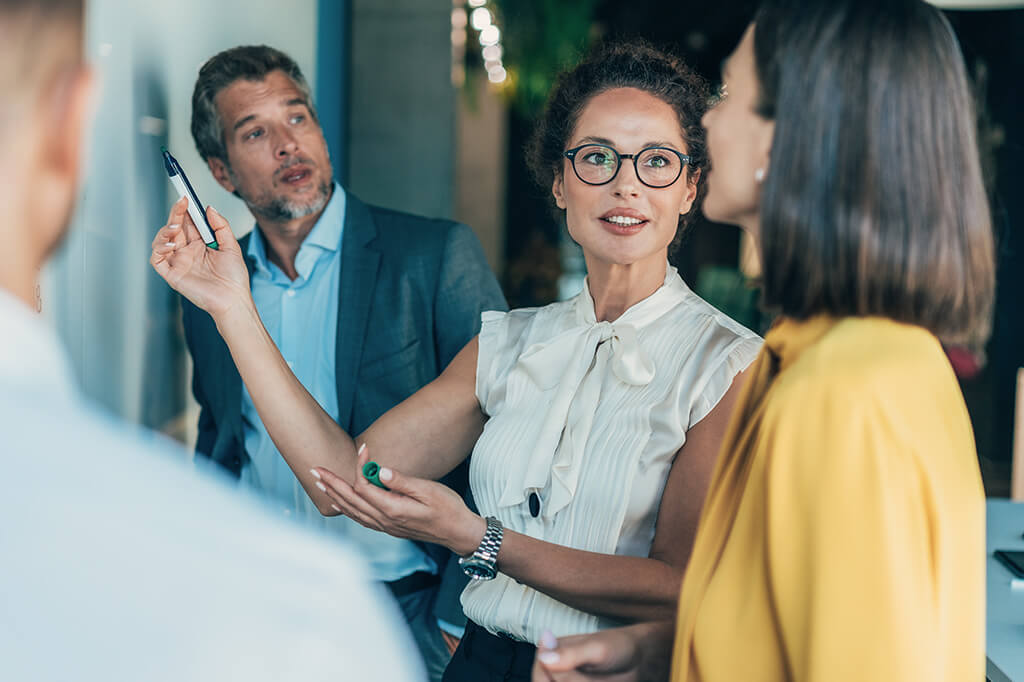 A woman in glasses and a white blouse gestures with a pen while leading a discussion at a whiteboard with colleagues. Two coworkers, a man in a suit and a woman in a yellow blouse, listen attentively during the meeting.