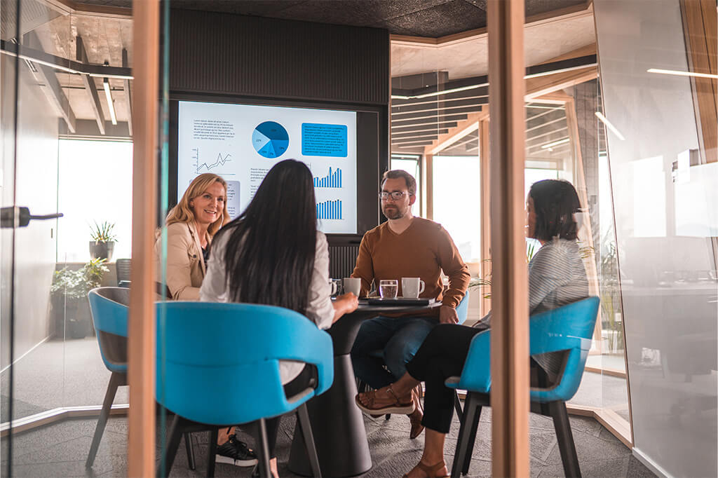 A group of four colleagues sit around a small conference table in a modern glass-walled office, reviewing charts and graphs displayed on a screen during a team meeting.