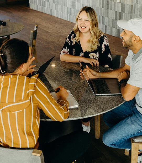 Three people sit together around a round table in a casual meeting. A woman in a yellow striped shirt takes notes on a laptop while another woman in a black floral dress smiles and speaks. A man wearing a white cap and gray shirt listens attentively with his laptop open. The setting appears to be a relaxed, modern workspace with warm lighting and wood flooring.