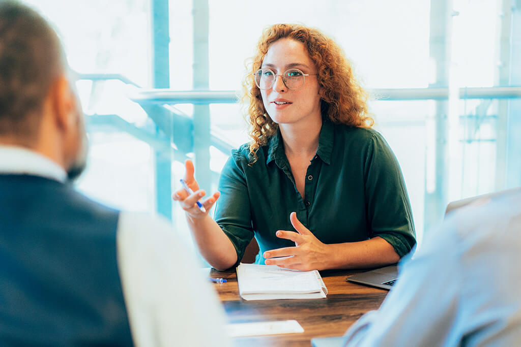 A woman with curly red hair and glasses speaks confidently during a meeting, gesturing with her hands while seated at a table with two colleagues in a bright, modern office.