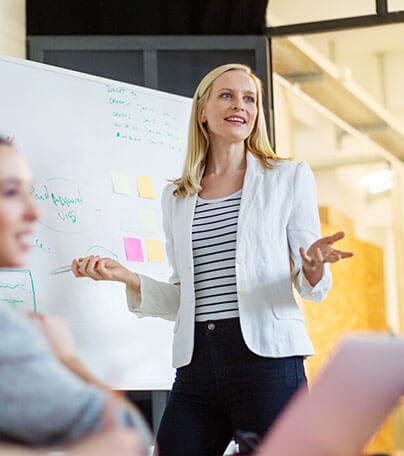 A woman in a white blazer and striped shirt stands in front of a whiteboard, smiling and speaking to colleagues during a presentation in a bright office.