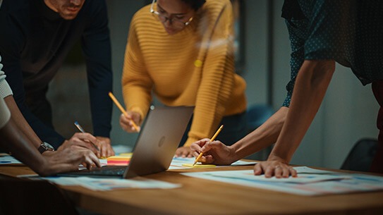 Group of people collaborating around a table, reviewing papers and sketches beside an open laptop in a modern office setting.